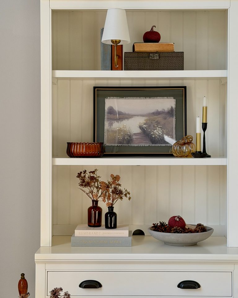 White shelves styled for fall with framed landscape art, flowers, books, and pumpkins.
