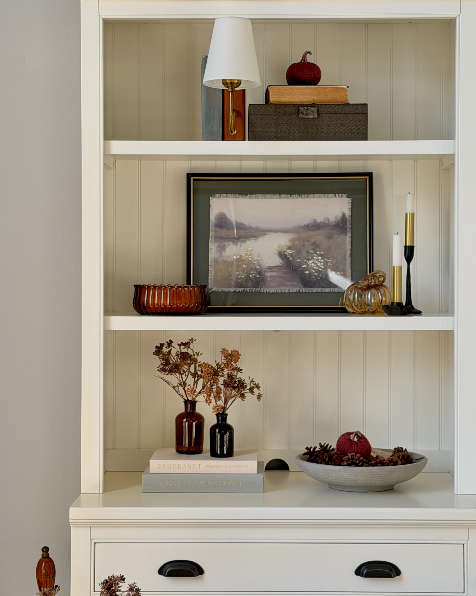 White shelves styled for fall with framed landscape art, flowers, books, and pumpkins.