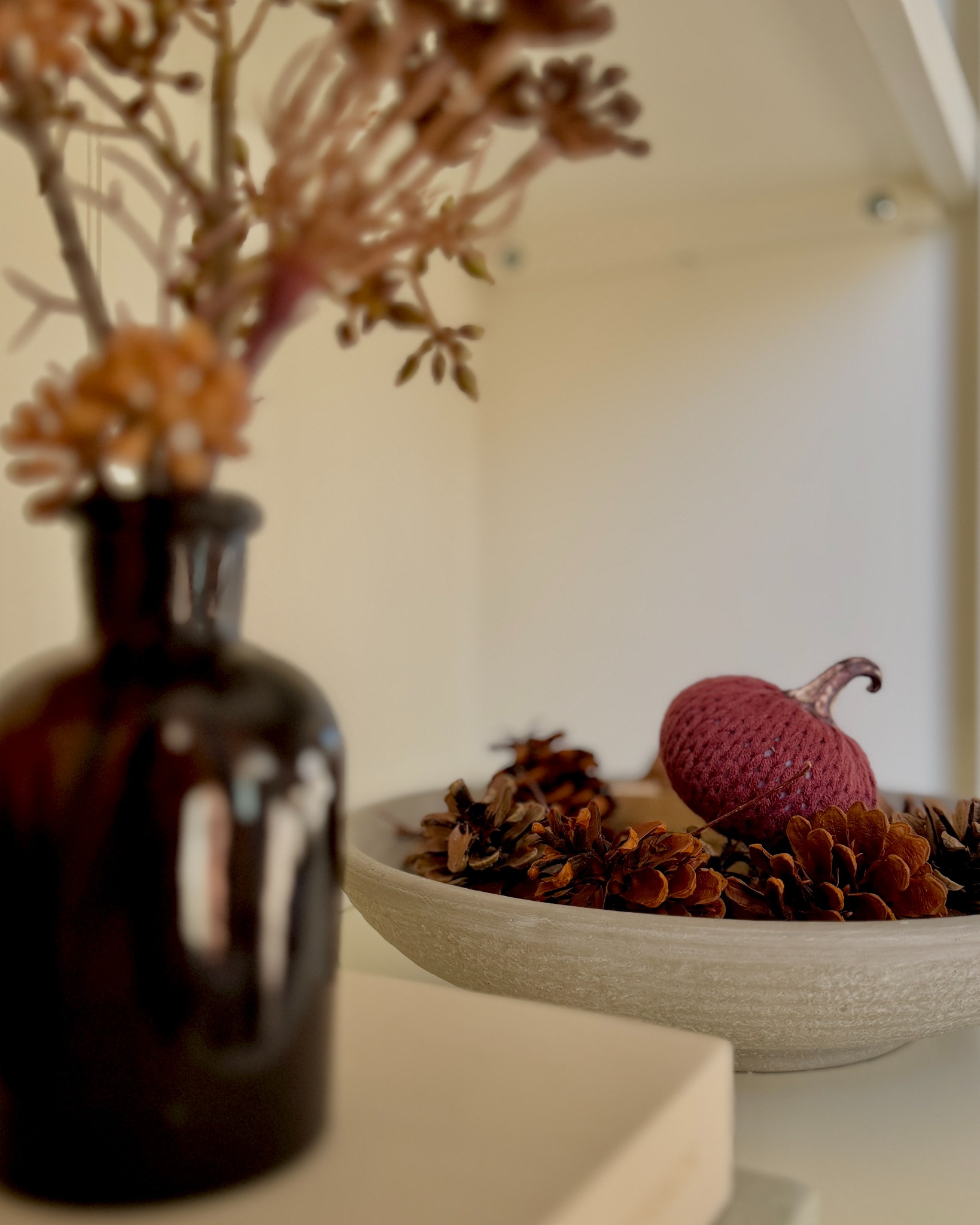 Fall flowers in an amber glass bottle beside a bowl of pinecones and a small pumpkin.